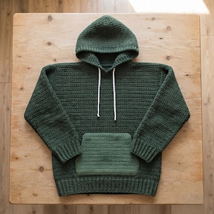 Peut inclure: Un sweat à capuche en crochet vert forêt foncé avec une capuche, des manches longues à poignets côtelés, un ourlet côtelé et une poche kangourou avant vert sauge plus clair. Deux fins cordons blancs pendent de l'encolure du vêtement.