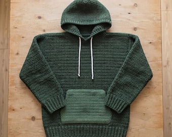 May include: A dark forest green crochet hoodie featuring a hood, long sleeves with ribbed cuffs, a ribbed hem, and a lighter sage green front kangaroo pocket. Two thin white drawstrings hang from the neckline of this textured yarn garment.