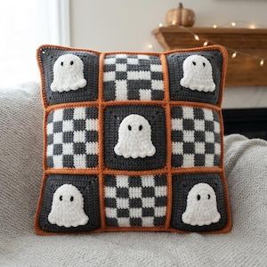 May include: A square crochet pillow featuring a grid of nine squares. Four squares contain white ghost appliques on dark gray backgrounds, while five squares feature a black and white checkered pattern. The entire grid is outlined in orange yarn.