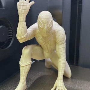 May include: A monochromatic, off-white 3D printed figurine of a superhero in a crouched, dynamic pose. The figure features a detailed web-like pattern across the entire suit, with one hand raised and the other touching the ground surface.