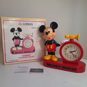 Puede incluir: Un reloj despertador parlante Lorus Mickey Mouse con base roja, botones amarillos y una bocina amarilla en la parte superior. La esfera es blanca con números negros. Al lado está la caja original con una ilustración y el texto Mickey Mouse.