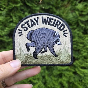 May include: A semi-circular embroidered patch featuring a dark gray raccoon walking through green grass. The background is a light beige color, and the text "STAY WEIRD" is arched in black thread at the top. The entire patch has a thick black border.