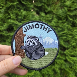 May include: A circular embroidered patch featuring a black and grey raccoon standing next to a brown tree trunk on a green grassy field. The background shows a light blue sky, a city skyline, and a mountain. The text JIMOTHY is arched at the top.