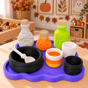 May include: A purple tray holding a collection of Halloween-themed items including a black cauldron with a white bone spoon, a white mummy bottle, an orange pumpkin jar, a bright green potion bottle, a candy corn cup, and a black bat cup.
