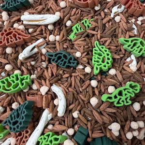 Peut inclure: Un mélange de remplissage pour bac sensoriel composé de grains de riz brun, de petites perles sphériques blanches, de pâtes blanches incurvées et de pâtes en forme de dinosaures vert foncé, vert clair et orange terre cuite.