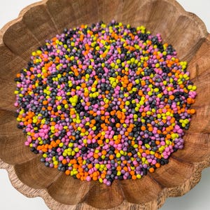 Peut inclure: Un bol en bois festonné rempli d'une collection dense de petites perles sphériques dans un mélange vibrant de couleurs noir mat, orange vif, jaune néon et rose magenta, créant un affichage texturé et coloré de minuscules billes rondes.
