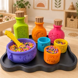 Könnte beinhalten: Ein Set aus bunten Silikon-Spielzeugen für sensorisches Spiel, bestehend aus einem lila Kessel mit gelbem Löffel, drei Flaschen mit Holzstopfen in Grün, Orange und Pink sowie drei kleinen Bechern in Blau, Orange und Gelb, gefüllt mit kleinen Perlen.