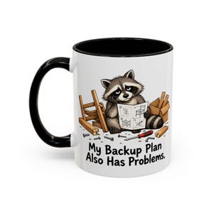 May include: A white ceramic mug with a black handle and interior, featuring a cartoon illustration of a raccoon looking at assembly instructions surrounded by wooden furniture parts, a screwdriver, and the text My Backup Plan Also Has Problems.