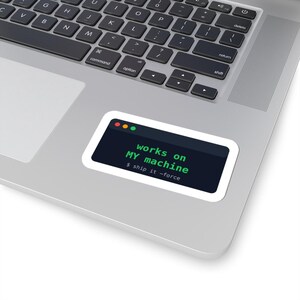 May include: A rectangular sticker with a white border applied to a silver laptop. The sticker features a dark interface window with red, yellow, and green dots, displaying the green text "works on MY machine" and "$ ship it -force".