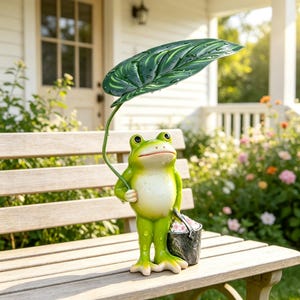 Può includere: Una statuetta di rana verde in piedi, che tiene una grande foglia verde scuro sopra la testa come un ombrello. La rana ha il ventre chiaro e porta nell'altra mano un piccolo secchiello grigio scuro pieno di fiori bianchi.