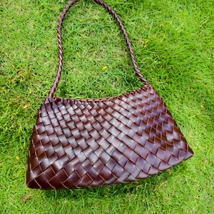 Puede incluir: Un bolso de mano de color marrón oscuro con un patrón trenzado similar a una cesta en toda la superficie exterior. El bolso tiene una sola correa de hombro fina y trenzada unida a las esquinas superiores, creando una forma rectangular.
