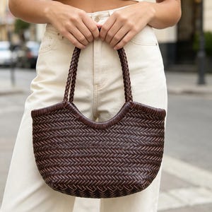 Puede incluir: Un bolso de mano trenzado de color marrón oscuro con una silueta en forma de media luna y un patrón de espiga ajustado en todo el cuerpo. El bolso incluye dos asas superiores trenzadas a juego fijadas al borde superior de la estructura.