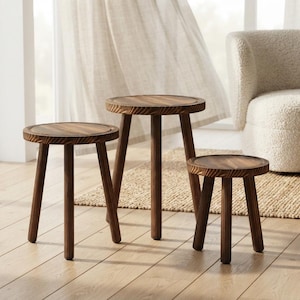 May include: A set of three round wooden side tables with dark brown wood grain tops and three cylindrical legs each. The tables vary in height, with the tallest positioned in the center and two shorter tables placed on either side of it.
