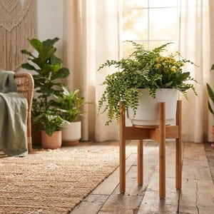 May include: A white cylindrical plant pot sits inside a light brown wooden stand with four legs. The pot contains a lush green fern and small yellow flowers. The stand is constructed from four vertical wooden pieces joined by a cross-shaped base.