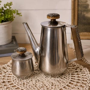 May include: A stainless steel coffee pot and a matching sugar bowl. Both pieces feature a polished silver finish, a tapered body, and a lid topped with a circular knob decorated with an intricate, dark, embossed floral pattern.