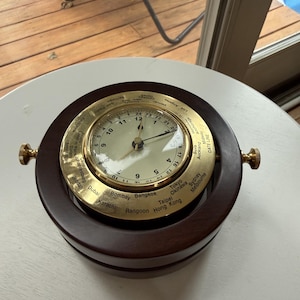 May include: A world time clock featuring a circular dark wood frame and a brass bezel. The clock face displays a 24-hour scale with central hands. The brass outer ring is engraved with various global city names and a date line for time zone tracking.