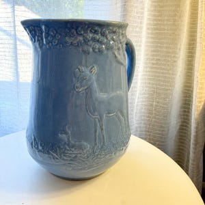 May include: A light blue ceramic pitcher featuring a raised relief design of a standing deer and a smaller fawn in a grassy field. The upper rim of the pitcher is decorated with a textured pattern of small circular shapes, and a handle is attached.