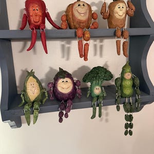 Può includere: Sette statuette di verdure realizzate a mano con volti sorridenti e arti pendenti, tra cui un peperone rosso, una zucca, un tubero, mais, una melanzana, broccoli e un baccello di piselli, disposte su due ripiani di una mensola blu.