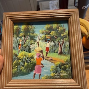 May include: A square framed painting depicting three figures walking through a lush green landscape with trees and a stream. The figures carry baskets on their heads, wearing colorful clothing including orange tops and pink or red skirts.