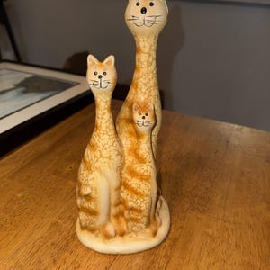 May include: A ceramic figurine featuring three cats of varying heights standing together on a circular base. The cats have a light cream base colour with orange tabby-style mottled markings, painted black eyes, noses, and whiskers on their faces.