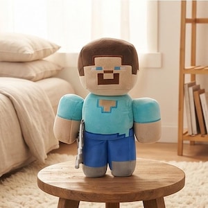 May include: A plush toy character featuring a blocky, pixelated design with a brown hair square head, light blue shirt, dark blue pants, and gray shoes. The figure has a tan face with blue eyes and a brown mouth, holding a small gray accessory.