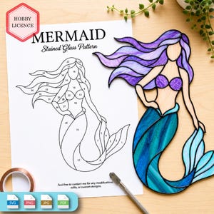 Puede incluir: Un atrapasoles de vidrieras con forma de sirena de cabello morado y cola azul, junto a un patrón de papel impreso etiquetado como MERMAID Stained Glass Pattern con secciones numeradas, cinta de cobre y una herramienta para cortar vidrio.