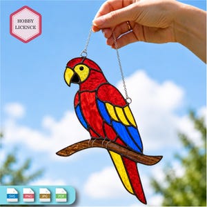 May include: A stained glass suncatcher shaped like a parrot with red, yellow, and blue glass segments, perched on a brown branch. The parrot has a yellow face and a black beak. A metal chain is attached to the top for hanging. The text HOBBY LICENCE is visible in a pink hexagon in the upper left corner.