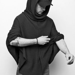 Könnte beinhalten: Ein schwarzer Kapuzenpullover mit einem Rollkragen und einer schwarzen Baseballkappe. Der Pullover wird von einer Person getragen und die Ärmel werden hochgezogen.