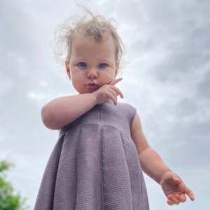 May include: A young child wearing a purple knitted dress, looking directly at the camera with a serious expression. The child's hair is windblown and the background is a cloudy sky.