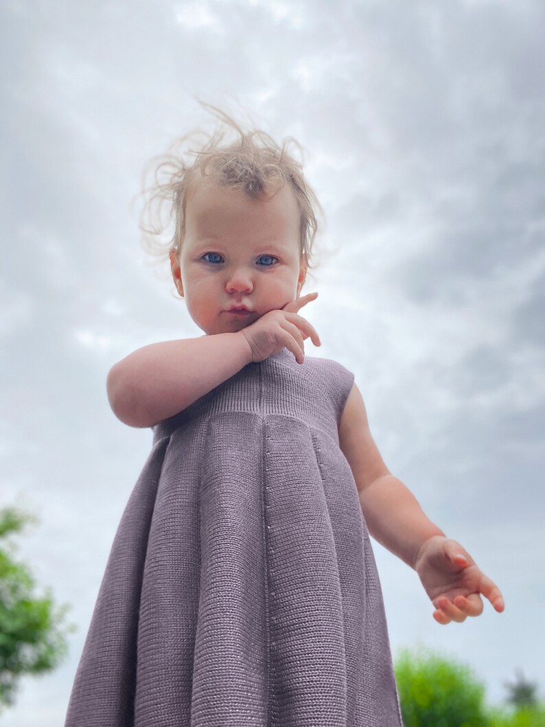 May include: A young child wearing a purple knitted dress, looking directly at the camera with a serious expression. The child's hair is windblown and the background is a cloudy sky.