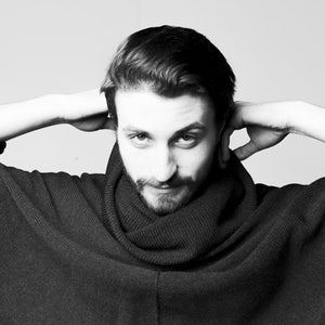 May include: A man wearing a black turtleneck sweater with a relaxed fit. He is looking directly at the camera with his hands behind his head.