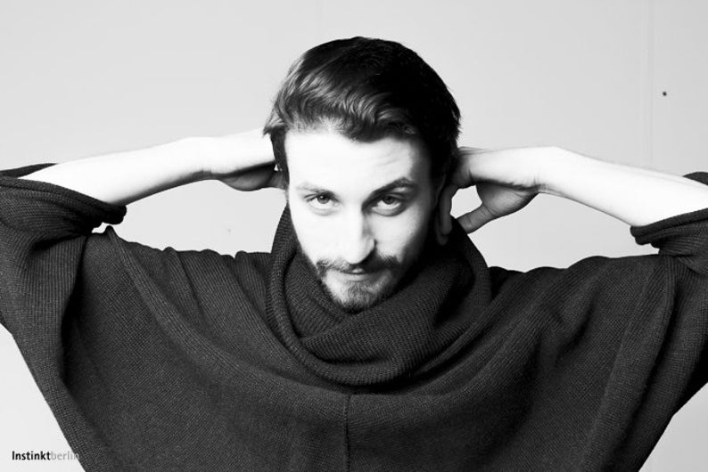 May include: A man wearing a black turtleneck sweater with a relaxed fit. He is looking directly at the camera with his hands behind his head.