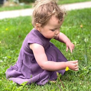 May include: A young child wearing a purple dress sits in a grassy field, looking down at a yellow dandelion flower.