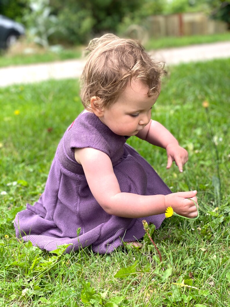 May include: A young child wearing a purple dress sits in a grassy field, looking down at a yellow dandelion flower.