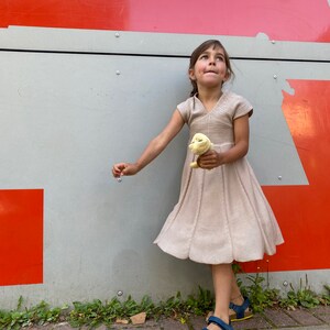 May include: A young girl in a beige dress and blue sandals stands in front of a red and gray wall. She is holding a yellow stuffed animal and looking up.