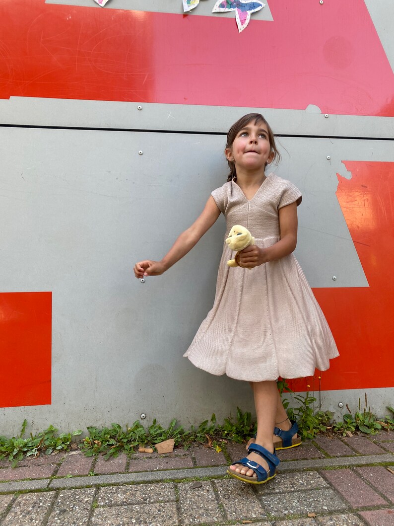 May include: A young girl in a beige dress and blue sandals stands in front of a red and gray wall. She is holding a yellow stuffed animal and looking up.