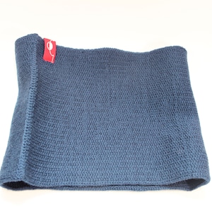 Puede incluir: Un cuello o bufanda de punto azul. El cuello está doblado por la mitad y tiene una etiqueta roja.