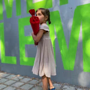 May include: A young person wearing a beige dress and blue sandals holds a red and white cone-shaped object. The person is standing in front of a green and gray wall with the word "LE" visible.