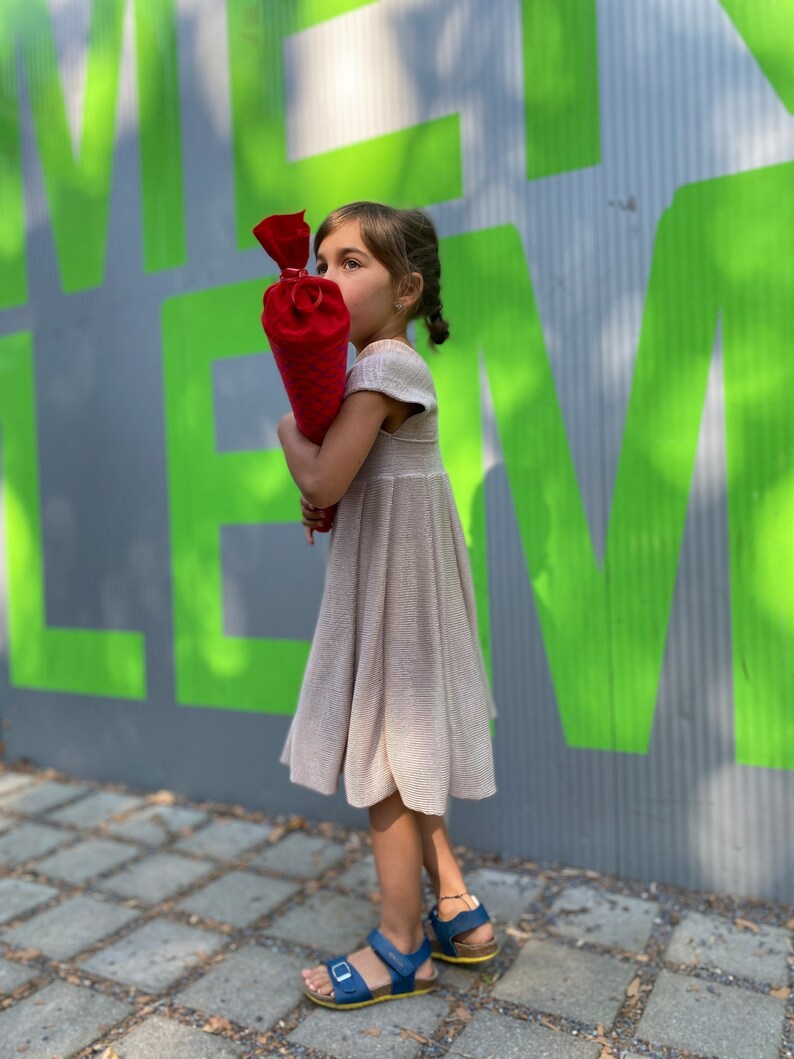 May include: A young person wearing a beige dress and blue sandals holds a red and white cone-shaped object. The person is standing in front of a green and gray wall with the word "LE" visible.