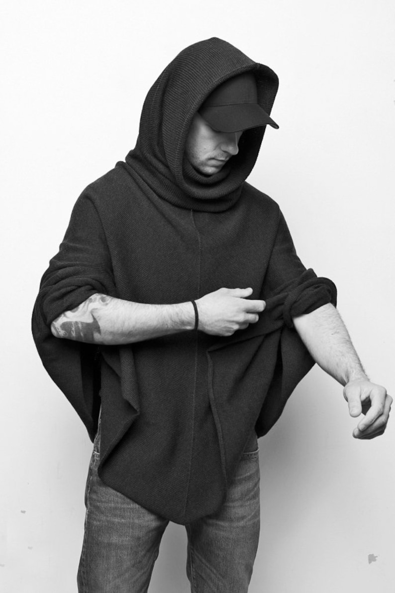 Könnte beinhalten: Ein schwarzer Kapuzenpullover mit einem Rollkragen und einer schwarzen Baseballkappe. Der Pullover wird von einer Person getragen und die Ärmel werden hochgezogen.