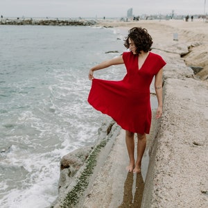 Eine junge Frau trägt das Kleid in Rot am Strand. Die Taille ist elastisch und wie bei einem Empire-Kleid hochgelegt. Der Rock mutet mit seinen Längsstreifen wie ein Faltenroch an.