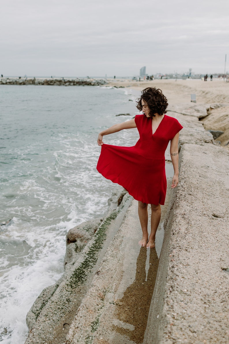 Eine junge Frau trägt das Kleid in Rot am Strand. Die Taille ist elastisch und wie bei einem Empire-Kleid hochgelegt. Der Rock mutet mit seinen Längsstreifen wie ein Faltenroch an.