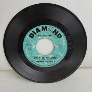 Peut inclure: Un disque vinyle noir 45 tours avec une étiquette centrale bleu clair portant le texte Diamond Records Inc, Don't Be Ashamed par Johnny Thunder, et un motif géométrique en diamant. L'étiquette inclut des crédits en texte noir.