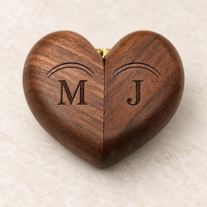 Puede incluir: Una caja de madera en forma de corazón hecha de madera de nogal marrón oscuro con un patrón de grano visible. La parte superior de la caja está grabada con las letras M y J, cada una rematada con un diseño de arco curvo. Las dos mitades del corazón se unen en el centro.