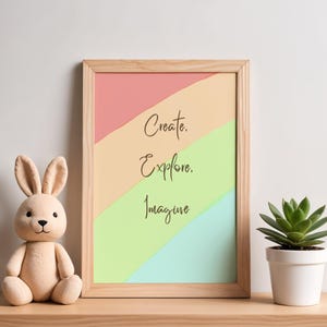 May include: A light wood framed print featuring diagonal stripes in pastel pink, peach, light green, and light blue. The text "Create. Explore. Imagine." is printed in black. A beige plush rabbit and a small green succulent in a white pot.