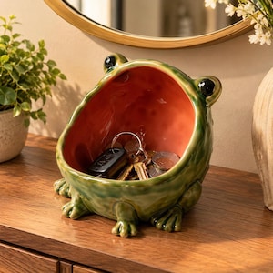 Może przedstawiać: Ceramiczna miska w kształcie żaby z błyszczącym zielonym wykończeniem na zewnątrz i gładkim pomarańczowym wnętrzem. Żaba ma dwoje czarnych oczu na górze i cztery płetwiaste stopy u podstawy. W misce znajduje się brelok z kluczami i monetami.