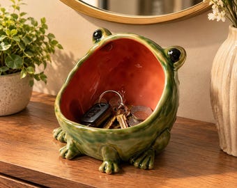 Puede incluir: Un cuenco de cerámica con forma de rana, exterior verde brillante e interior naranja liso. La rana tiene dos ojos negros en la parte superior y cuatro patas palmeadas en la base. El cuenco contiene un llavero con varias llaves y monedas.