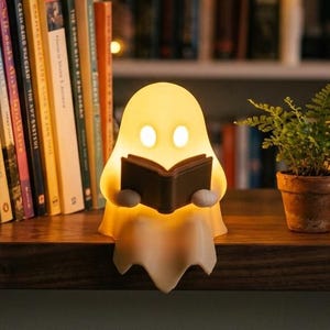 May include: A glowing, off-white, ghost-shaped lamp sitting on a shelf. The ghost figure has two oval-shaped illuminated eyes and is holding a small, dark brown, open book in its hands. The lamp emits a soft, warm light from within.