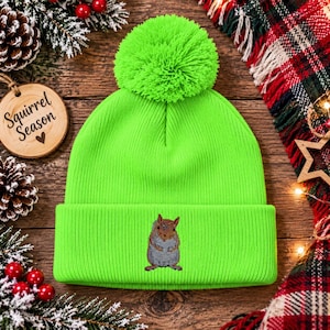 May include: A bright neon green knit beanie featuring a large matching pom-pom on top and a folded cuff. The center of the cuff displays a detailed embroidered graphic of a small brown squirrel standing upright in a neutral pose.