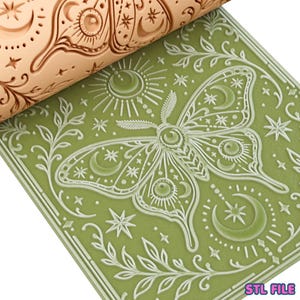 May include: A light tan cylindrical rolling pin with a deeply engraved moth, moon, and star pattern, positioned above a flat, sage green square surface that displays the same intricate embossed design of a large moth surrounded by celestial motifs.